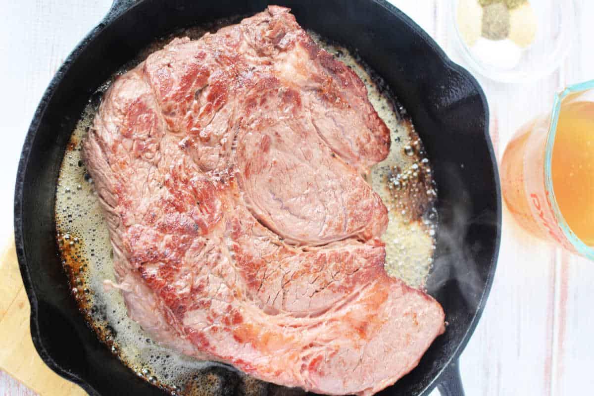 A large boneless chuck roast sizzles in a cast iron skillet, with seasonings and broth beside it.