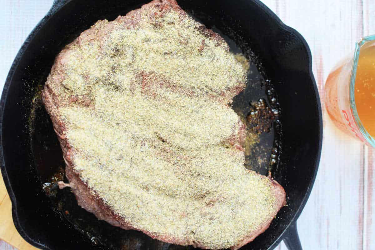 A seasoned boneless beef cut sizzles in a cast-iron skillet, resting enticingly beside a measuring cup filled with aromatic liquid.