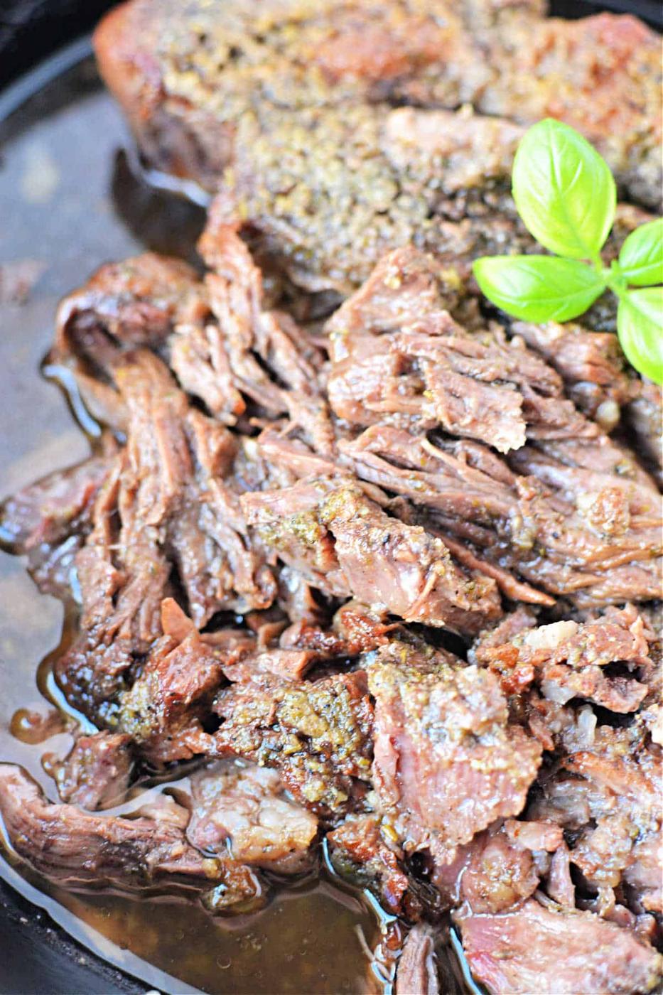 Close-up of shredded boneless chuck roast in sauce, adorned with a garnish of fresh green basil leaves.