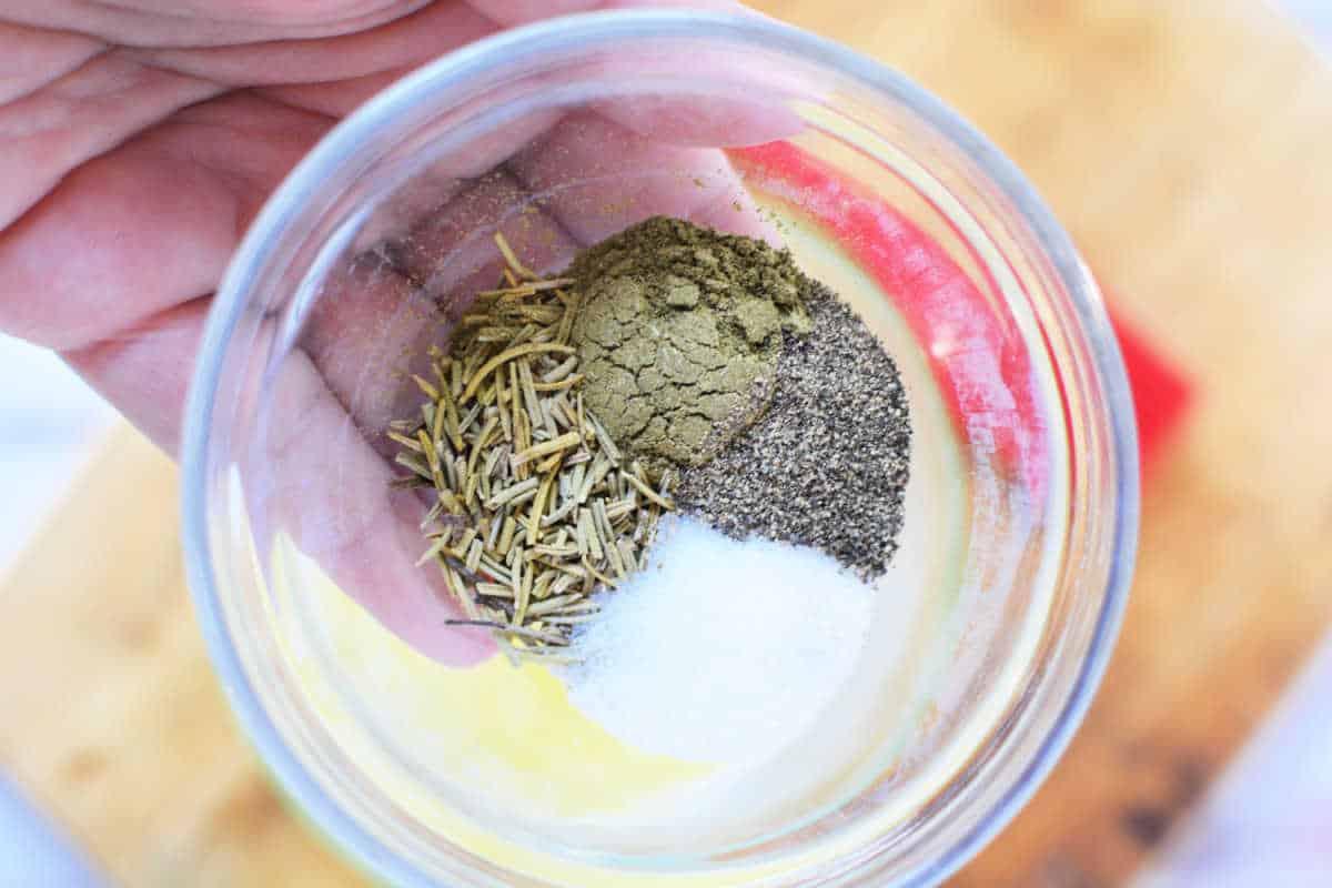 A hand holding a clear glass bowl containing a mix of dried rosemary, ground black pepper, salt, and another greenish-brown spice. Perfect for seasoning your Boneless Rib Roast Recipe. A wooden cutting board is visible in the background.