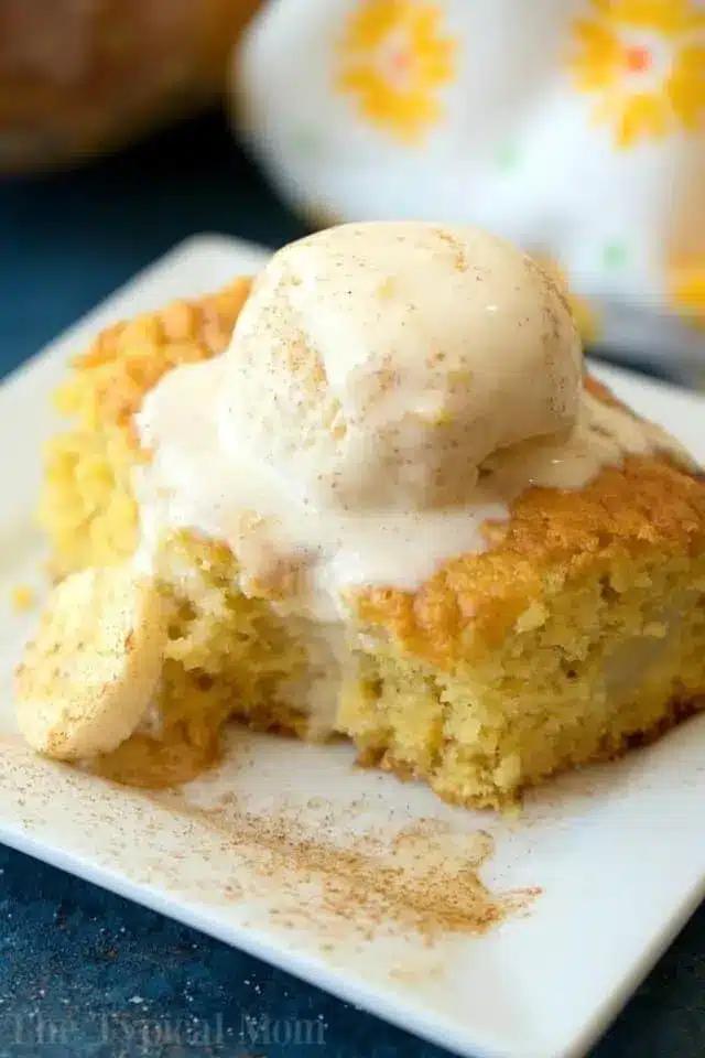 A slice of cake, made using clever box cake mix hacks, is topped with a scoop of vanilla ice cream on a white square plate. The cake boasts a light golden color, with a small cookie on the side. Cinnamon graces the top and surrounds the dessert, set against a backdrop of patterned cloth.