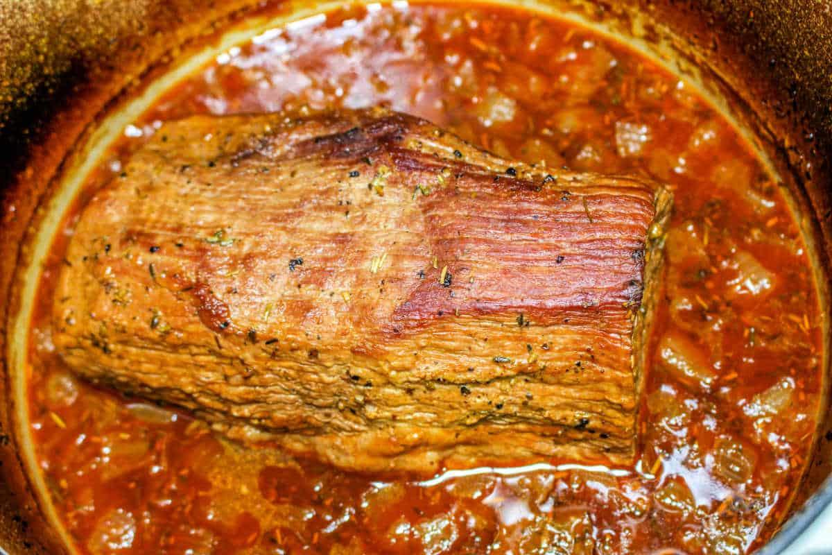 A succulent chuck roast braised to perfection in a pan, enveloped in a rich, brown sauce infused with red wine and adorned with visible herbs and spices.