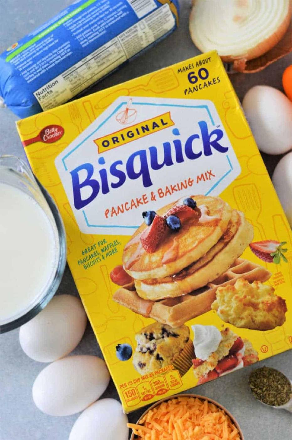 Box of Bisquick mix surrounded by eggs, milk, shredded cheese, an onion, and a roll of sausage—everything you need for a delicious Breakfast Casserole with Bisquick.