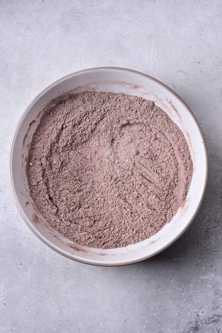 A bowl of mixed cocoa powder, perfect for whipping up a delightful brownie bread, sits gracefully in a white bowl on a textured gray surface.