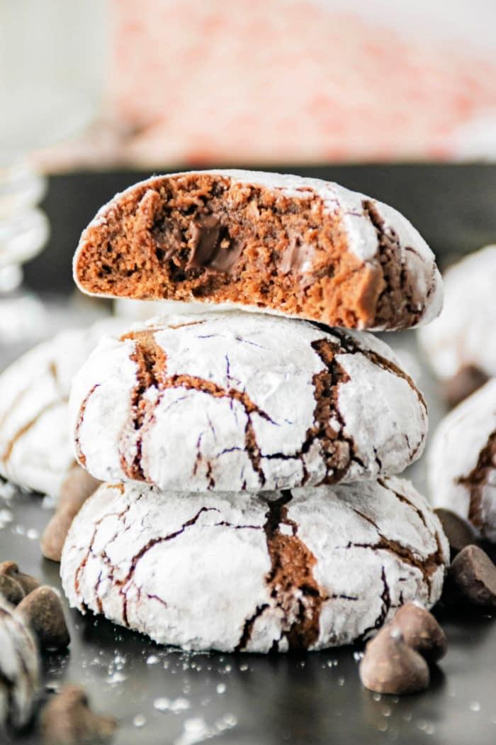 Three brownie mix crinkle cookies are stacked on a dark surface, with the top cookie broken in half to reveal the gooey chocolate inside. The cookies are dusted with powdered sugar, and chocolate chips are scattered around them.