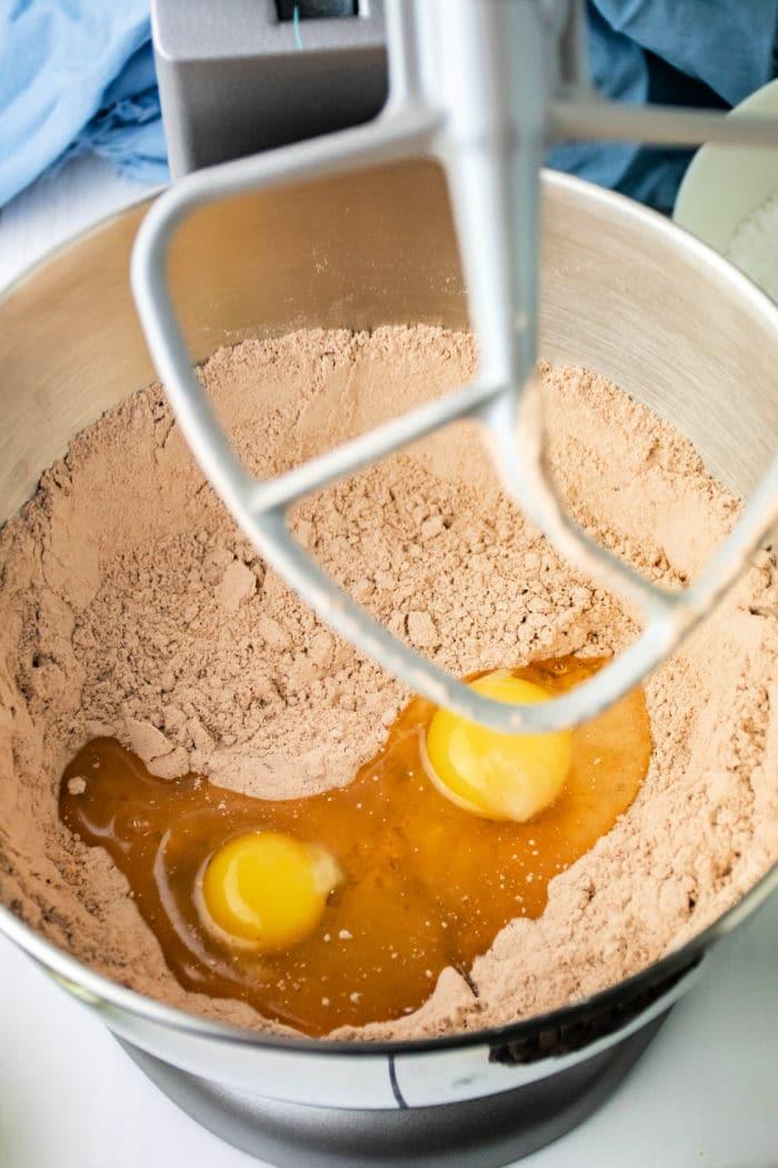 A mixing bowl filled with brown powder, likely brownie mix for crinkle cookies, and two cracked eggs. A metal stand mixer paddle is partially visible above the mixture. A blue cloth and other kitchen items are in the background.