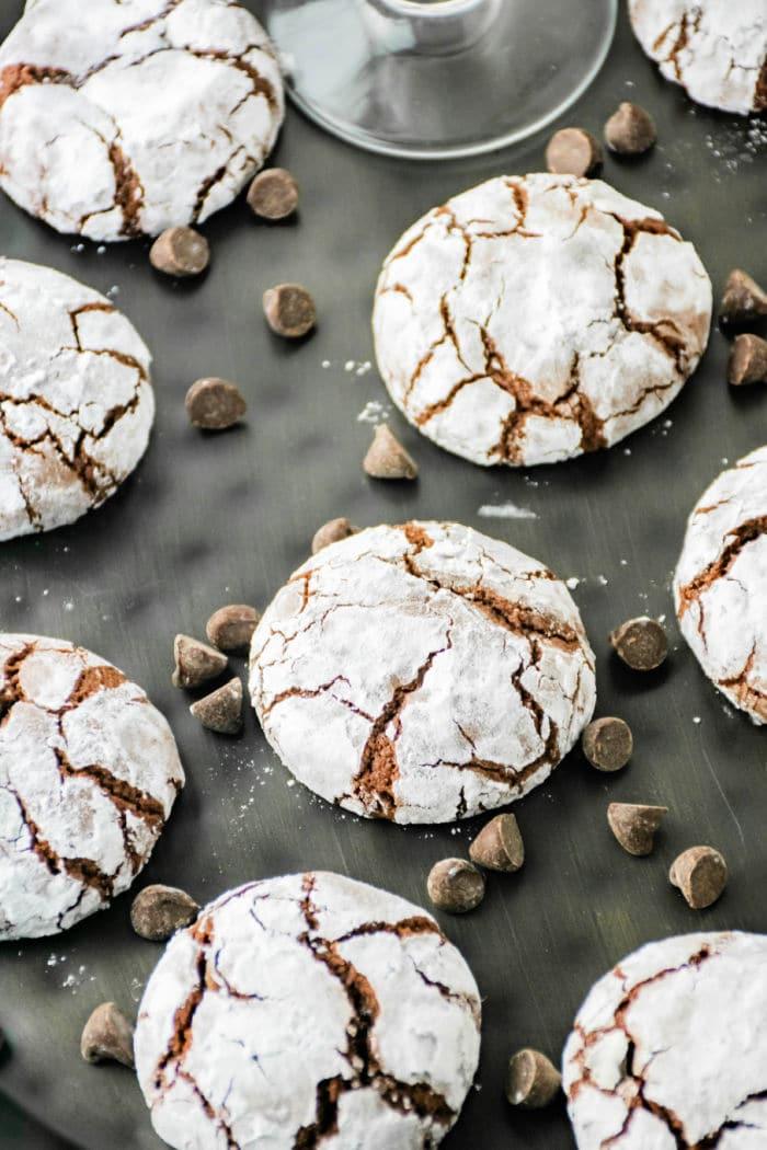 Crinkle cookies, crafted with brownie mix and dusted with powdered sugar, are arranged on a dark surface. Chocolate chips are scattered among the cookies, enhancing their richly cracked appearance.