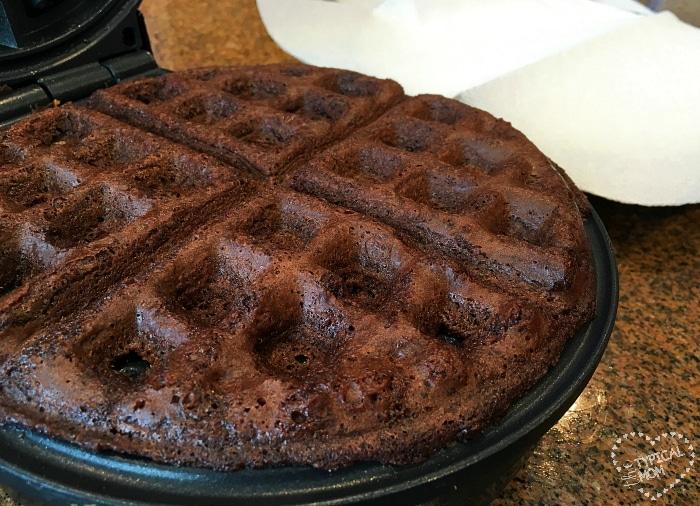 A delicious chocolate waffle, reminiscent of brownie waffles, sits enticingly inside a waffle maker, partially opened on the countertop.