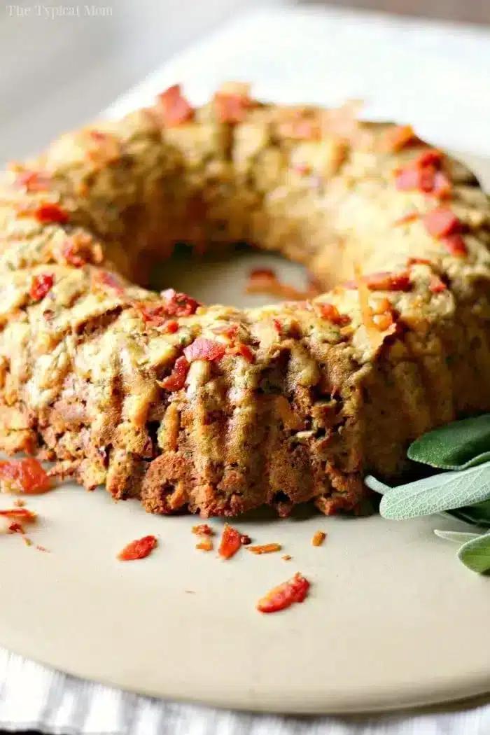 A savory bundt cake, shaped perfectly by the bundt pan, boasts a golden-brown crust and is topped with crispy bacon bits. Fresh sage leaves are artfully placed beside it on a beige surface, enhancing its rustic charm.