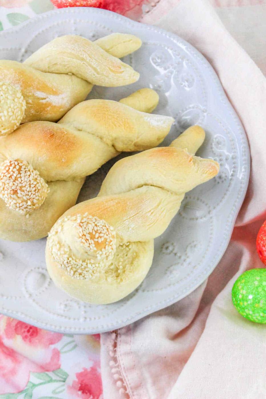 Twisted breadsticks with sesame seeds on a white plate, surrounded by colorful eggs and a floral tablecloth, offer a festive touch to your Bunny Bread recipe presentation.