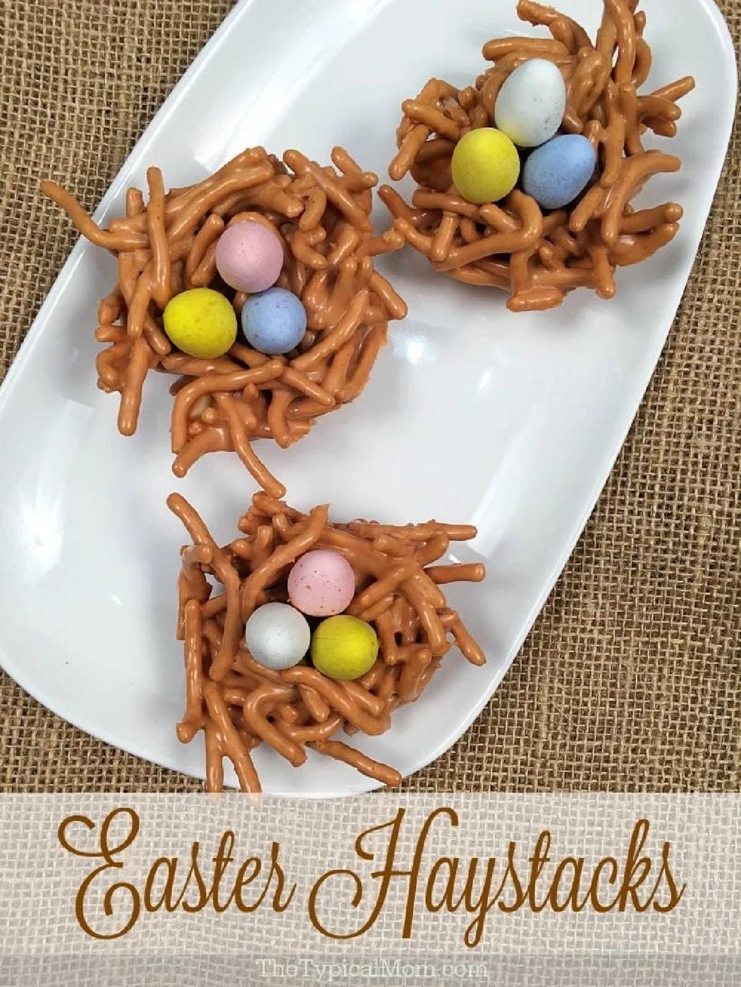 Three butterscotch haystacks shaped into edible nests, filled with pastel candy eggs on a white plate, set atop burlap fabric.