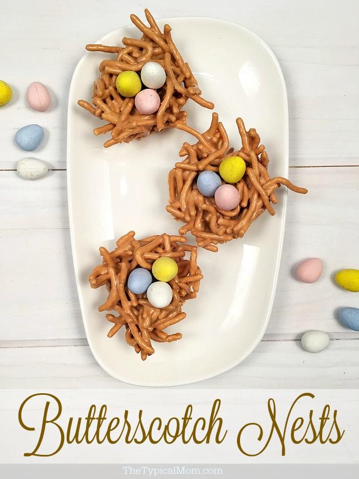 A white plate displays three delightful butterscotch haystacks crafted from pretzel sticks and butterscotch chips. Each nest cradles small, colorful candy eggs. The text Butterscotch Nests graces the bottom of the image.