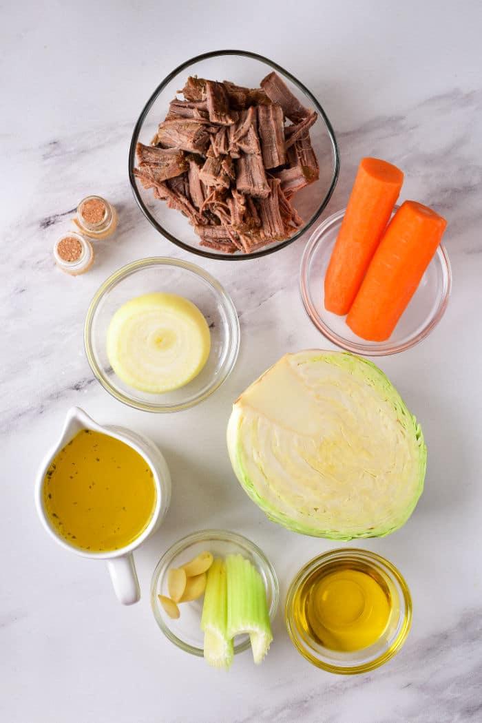 On a marble surface lie the ingredients for a delicious cabbage stew recipe: shredded beef, carrots, onion, cabbage, broth, garlic, celery, olive oil, and spices.