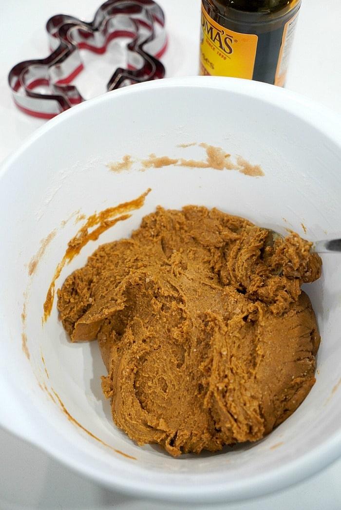 A white bowl contains rich gingerbread dough with a spoon sticking out, hinting at delicious gingerbread cookies to come. A bottle and cookie cutters are thoughtfully placed in the background, ready for action.