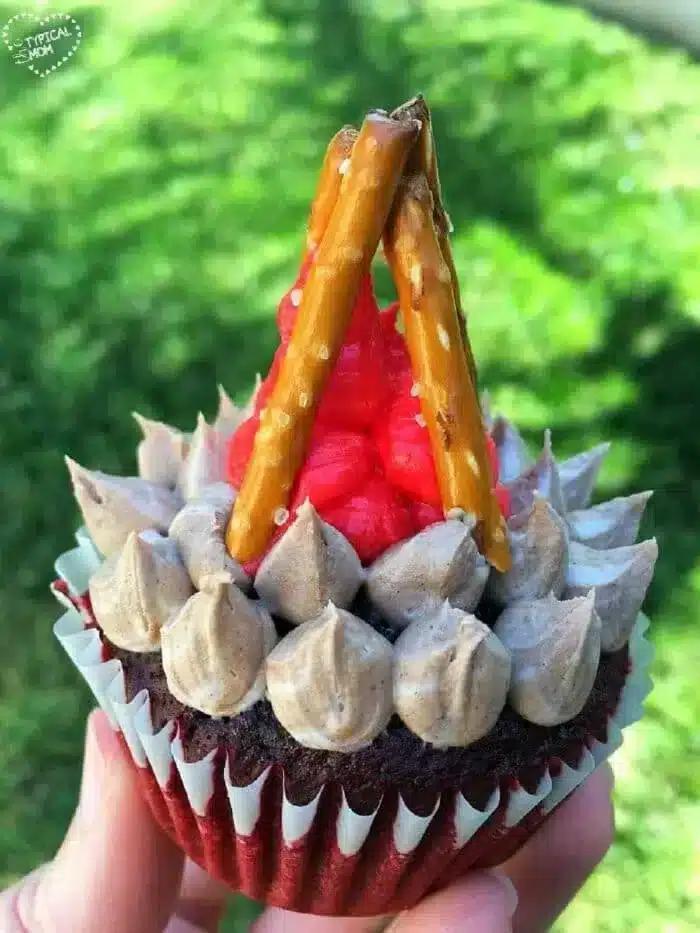 A Camping Cupcake masterfully decorated to resemble a campfire, with red frosting flames and pretzel stick logs. The chocolate base is topped with tan frosting peaks, all nestled in a hand against a grassy backdrop.