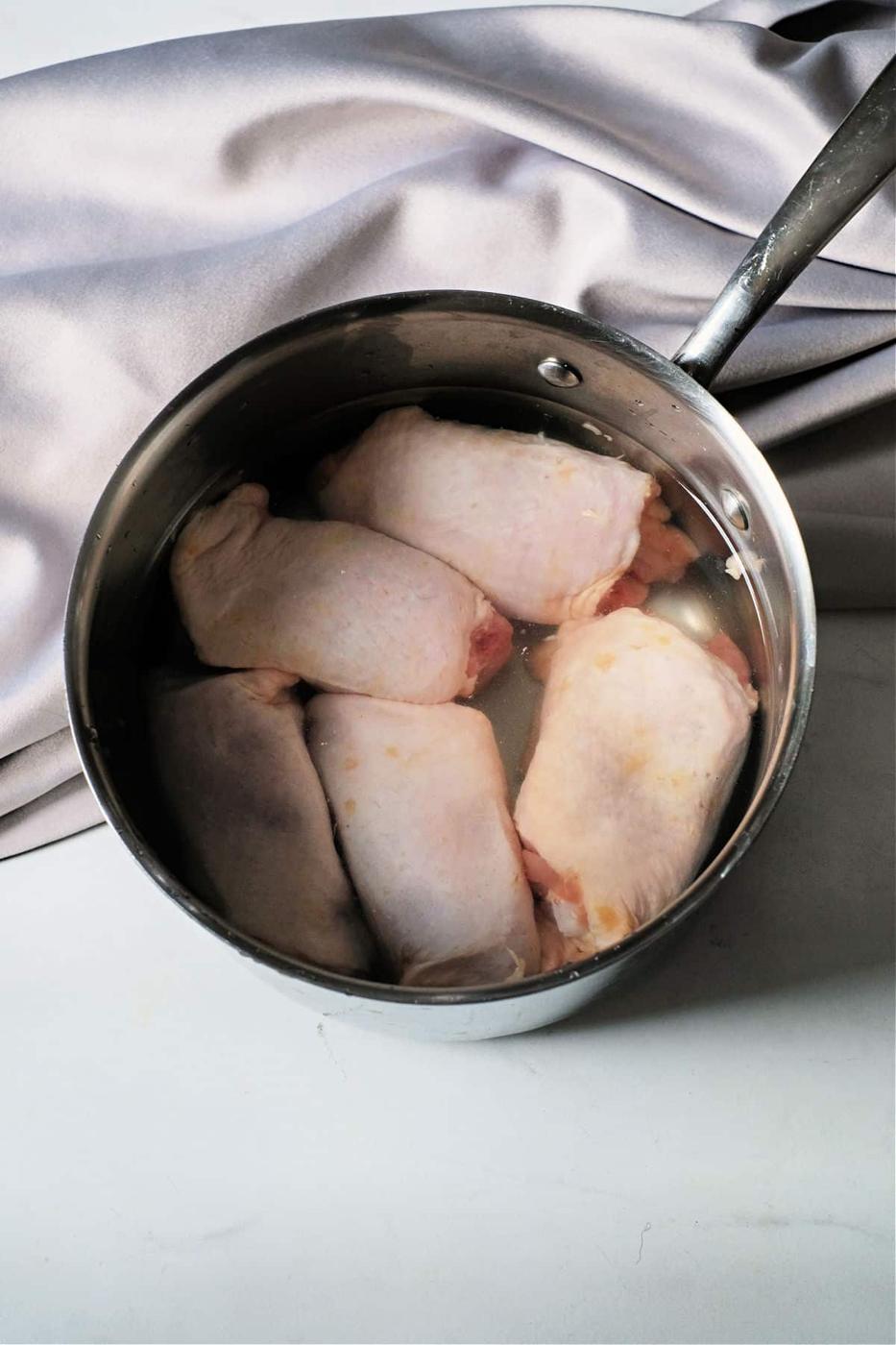 frozen chicken thighs rest in a metal pot filled with water on a marble surface, set against a silver cloth backdropâa perfect start for those wondering, Can you boil frozen chicken?.