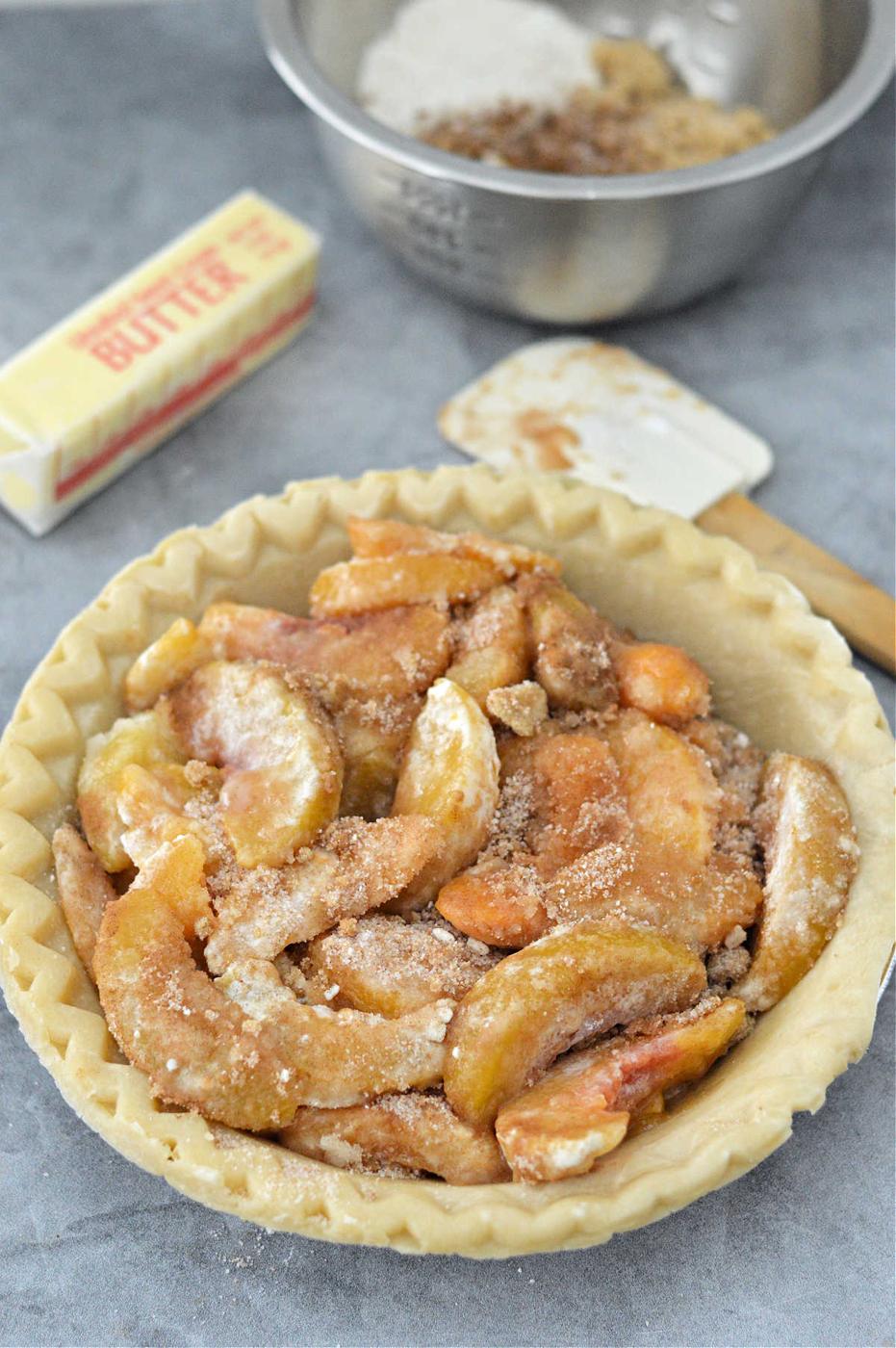Unbaked peach pie with crumb topping sits invitingly next to a stick of butter, flour, and a metal mixing bowl, reminiscent of preparing a delightful canned peach pie.