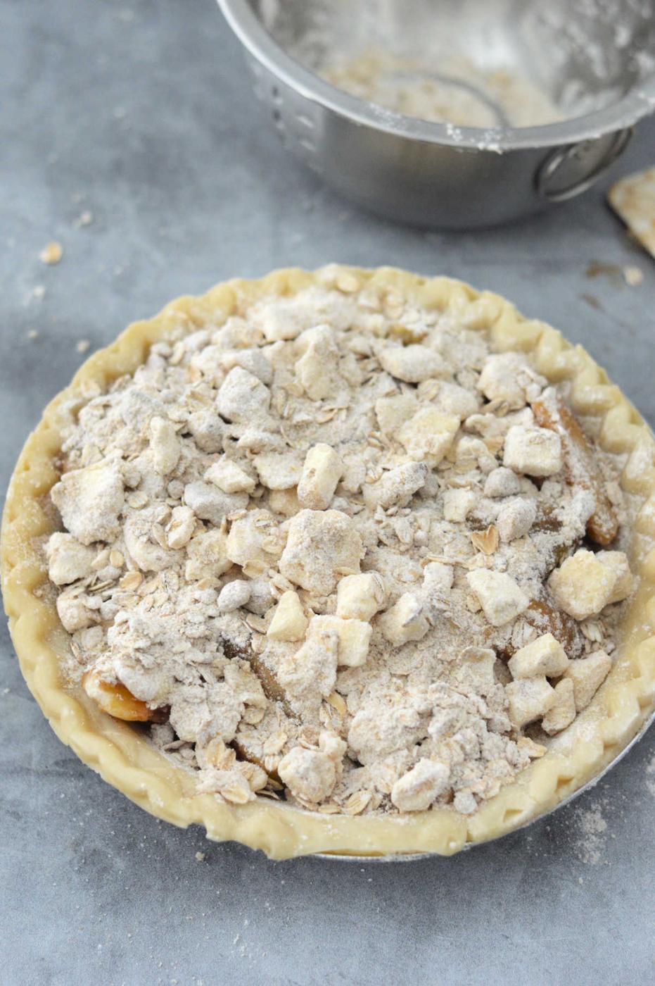 An unbaked peach pie with crumb topping sits enticingly on a gray countertop, while a metal bowl in the background hints at the possibility of crafting a delightful canned peach pie next.