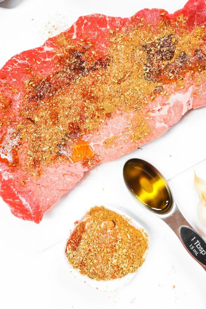 Raw marinated flank steak with seasoning, a tablespoon of oil, and a garlic clove on a white surface—perfect for slow-cooking into tender carne asada in your crock pot.