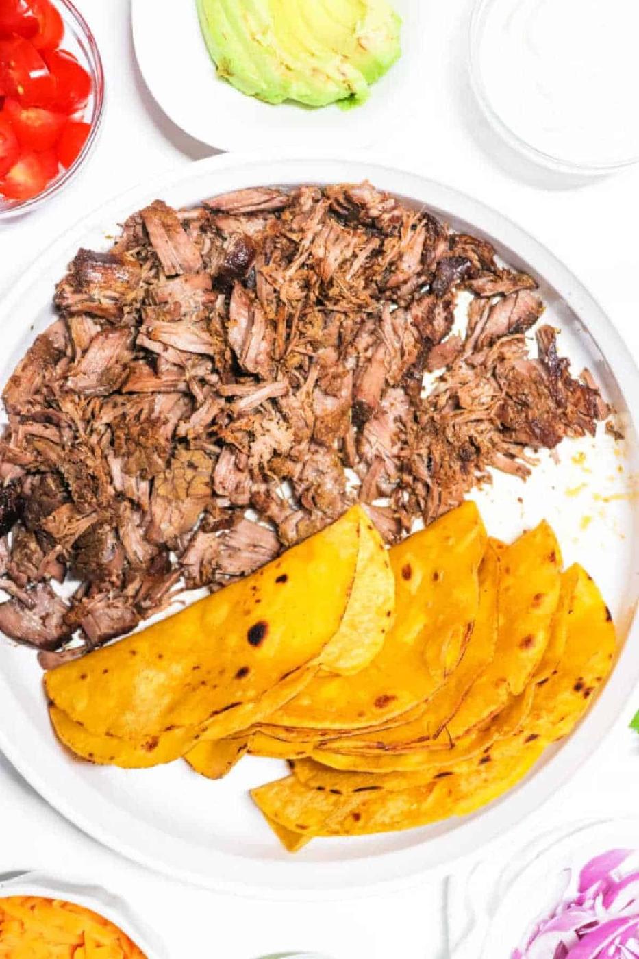 Shredded beef, slow-cooked like Carne Asada in a Crock Pot, and three yellow corn tortillas on a white plate, surrounded by taco toppings.