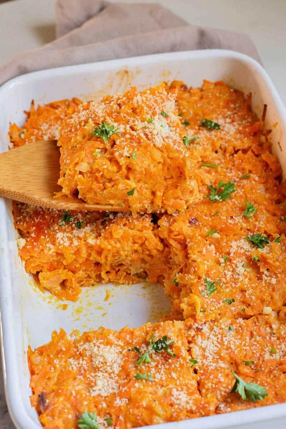 Cheesy carrot casserole in a white baking dish, topped with parsley, with a wooden spatula serving a piece.