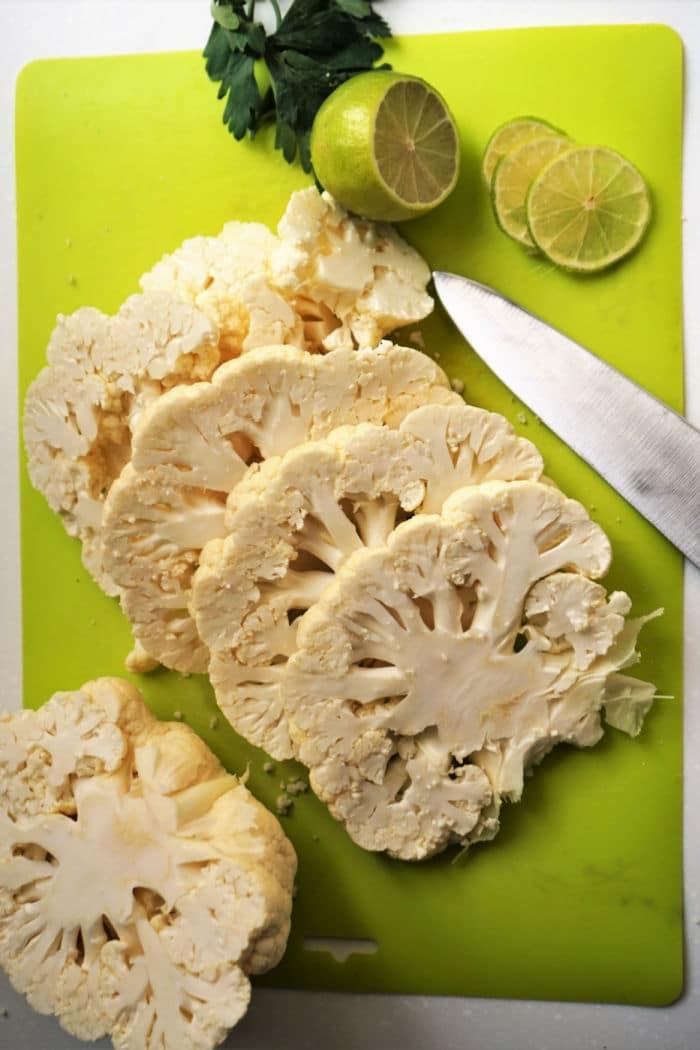 Sliced cauliflower, lime, and a knife on a green cutting board with parsley in the background—perfect ingredients for making cauliflower steak in the air fryer.