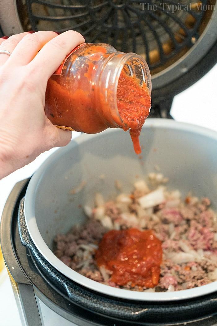 A person artfully pours spaghetti sauce over ground meat and onions in an open pressure cooker, preparing a savory base for cheesy Ninja Foodi spaghetti.