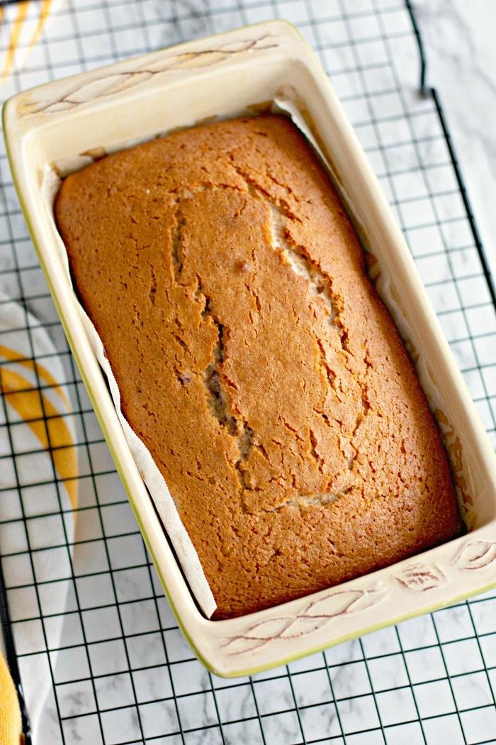 A freshly baked loaf of Cherry Bread in a rectangular ceramic pan rests on a cooling rack, its sweet aroma filling the air.