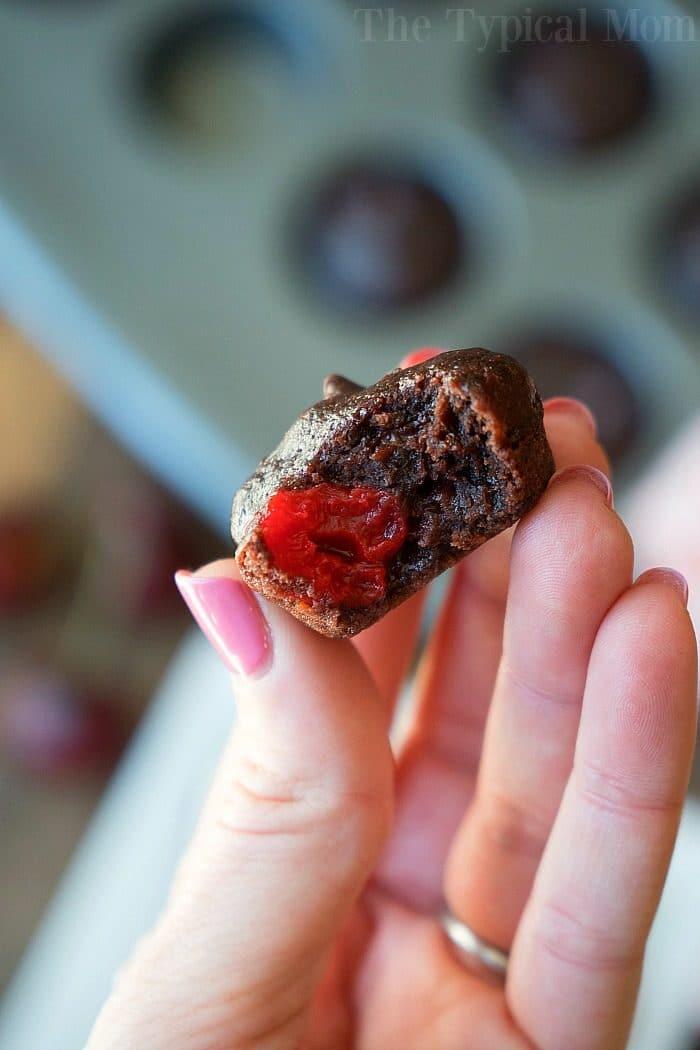 A close-up of a hand holding a small chocolate dessert with a bite taken out, revealing a bright red cherry filling inside. The background shows a blurred muffin tray filled with these delicious cherry brownies.
