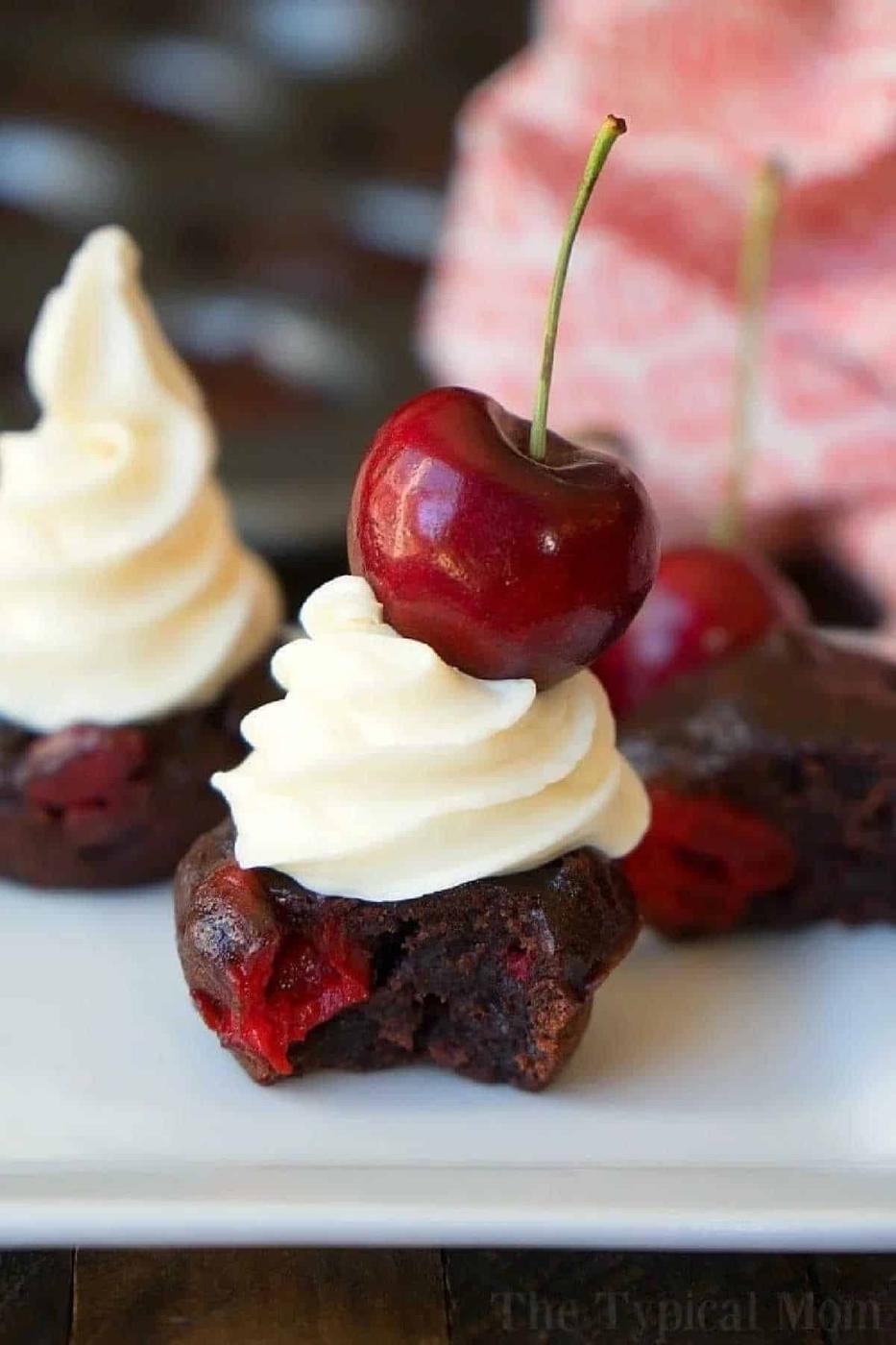 A decadent cherry brownie topped with smooth white frosting and a cherry, with a bite taken out to reveal its rich chocolatey center.