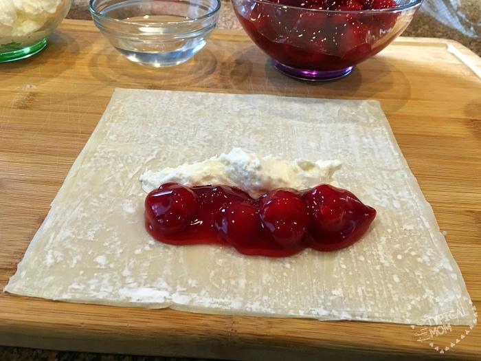 Square pastry sheet topped with cherry pie filling and cream cheese on a wooden board, reminiscent of cherry cheesecake egg rolls, with a bowl of cherry filling and water in the background.