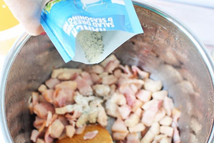Pouring seasoning into a pot of chicken crack soup with chopped bacon, stirring it all together with a wooden spoon.