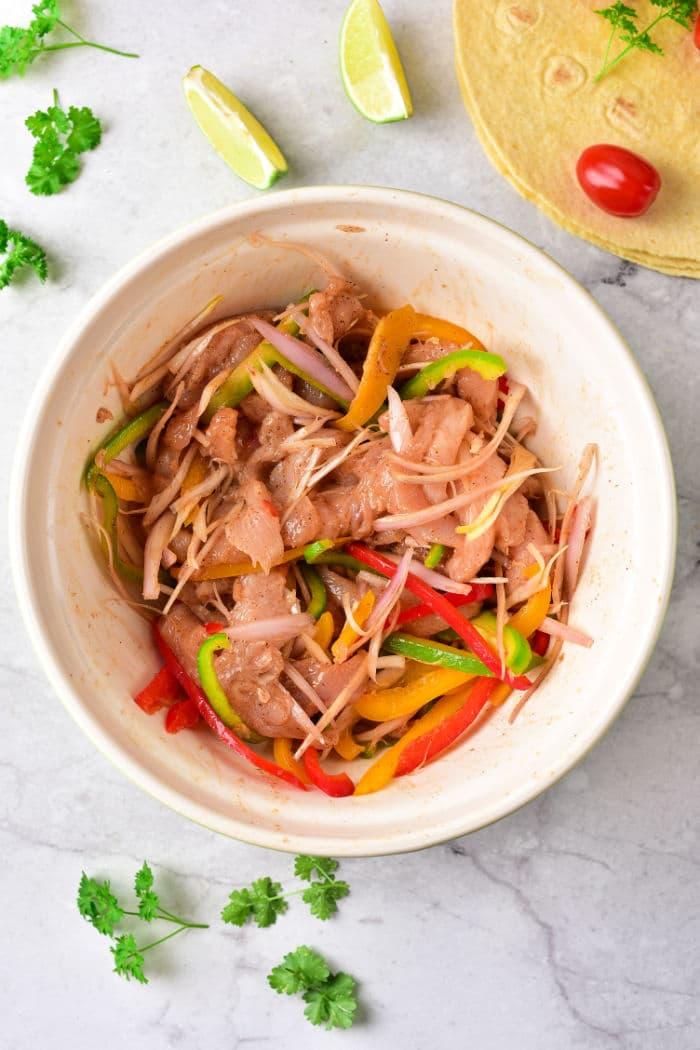 A bowl of marinated chicken with sliced bell peppers and onions, reminiscent of chicken fajita tacos, is surrounded by lime wedges and parsley for a vibrant presentation.