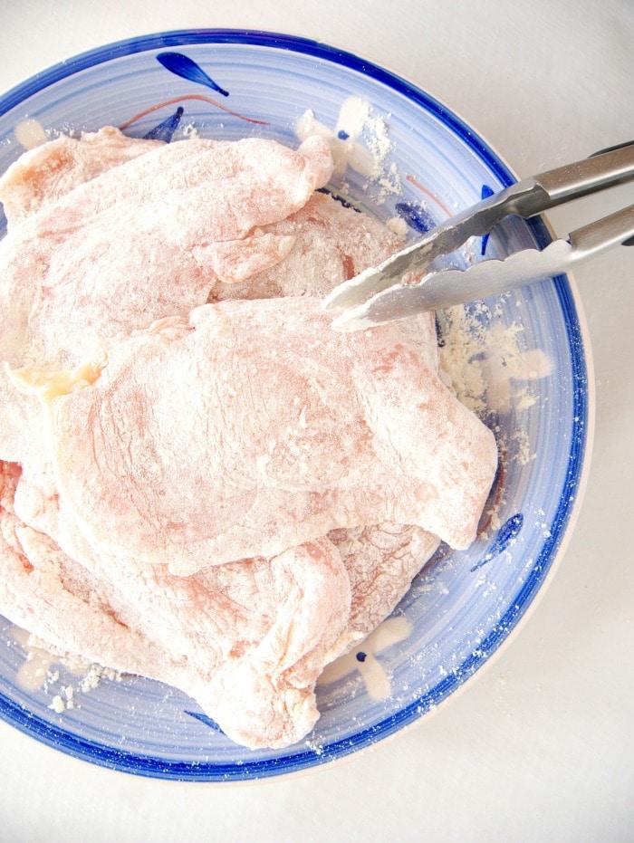 Raw chicken cutlet pieces, perfect for creating a quick chicken marsala, coated in flour on a blue plate with metal tongs.