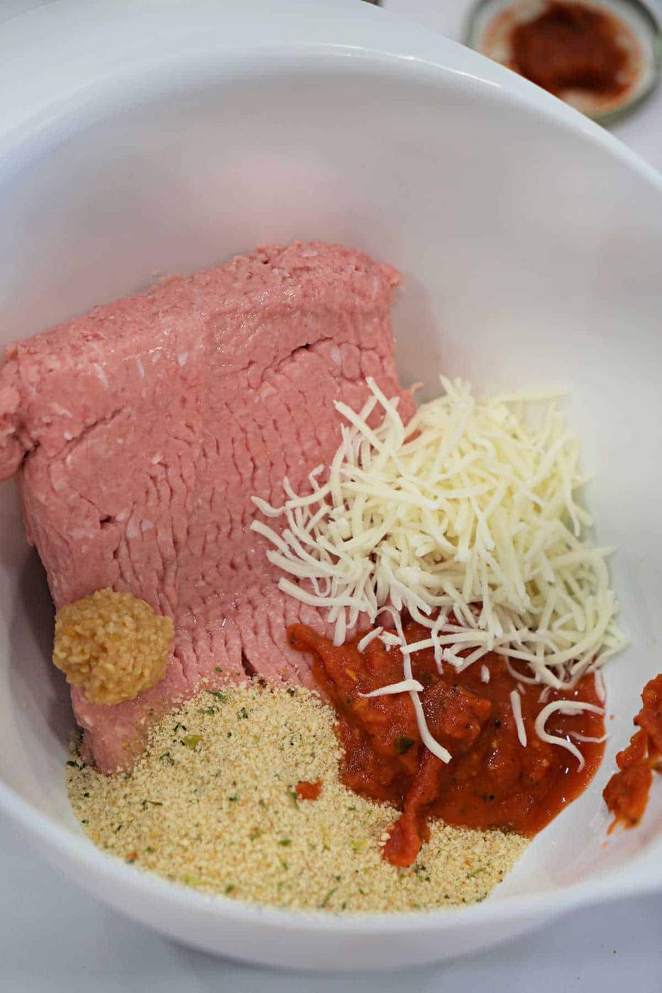 Ground meat, shredded cheese, breadcrumbs, minced garlic, and tomato sauce in a white mixing bowl—perfect for preparing a flavorful Chicken Parmesan Meatloaf.