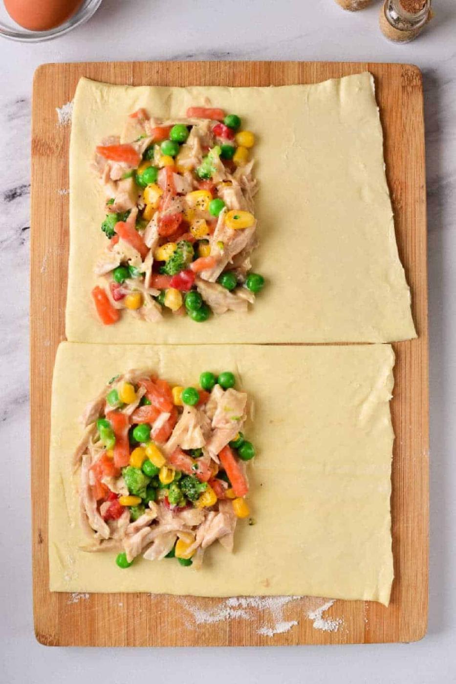 Two chicken pot pie puff pastry rectangles rest on a wooden board, each topped with a savory mix of vegetables and shredded chicken filling.