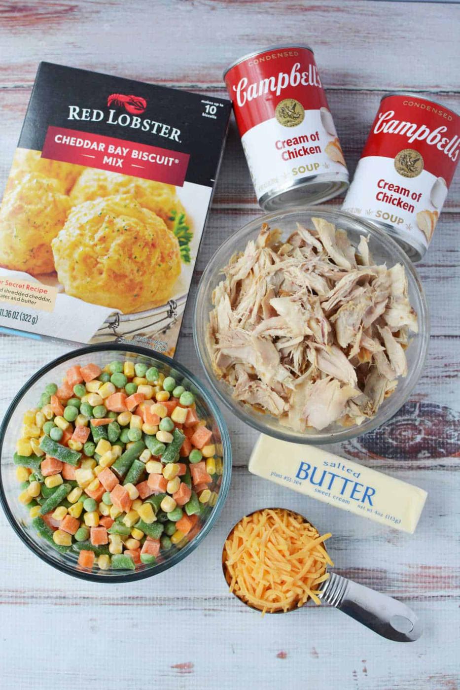 Ingredients for chicken casserole: biscuit mix or Red Lobster Biscuits, soup cans, shredded chicken, mixed veggies, butter, and shredded cheese for a Chicken Pot Pie-inspired dish.