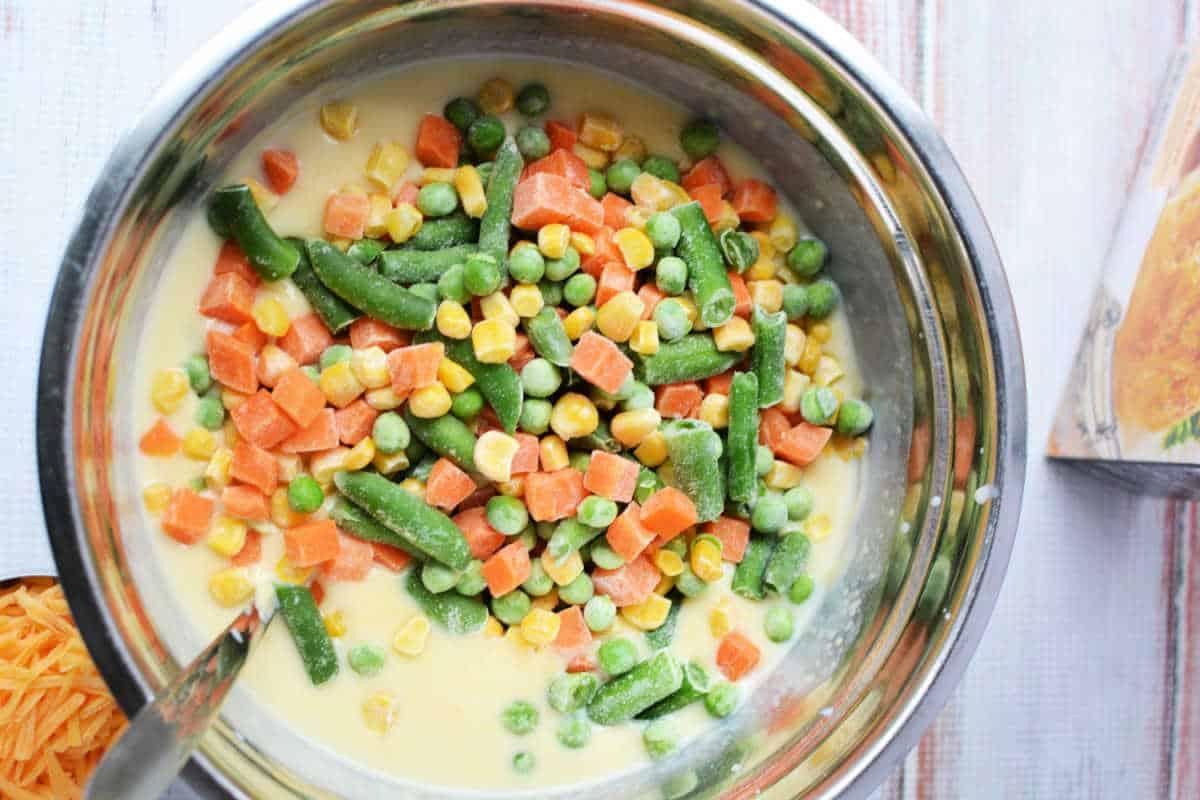 A bowl of mixed frozen vegetables in a creamy yellow sauce, reminiscent of Chicken Pot Pie filling, ready to be stirred.