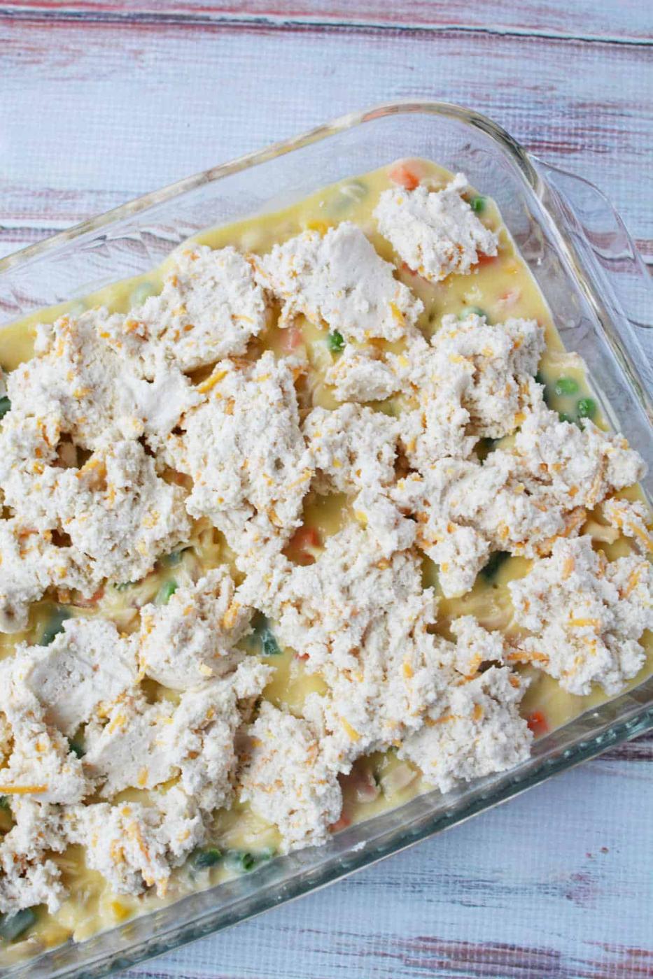 Unbaked chicken pot pie casserole topped with biscuits sits in a glass dish on a white wooden surface.