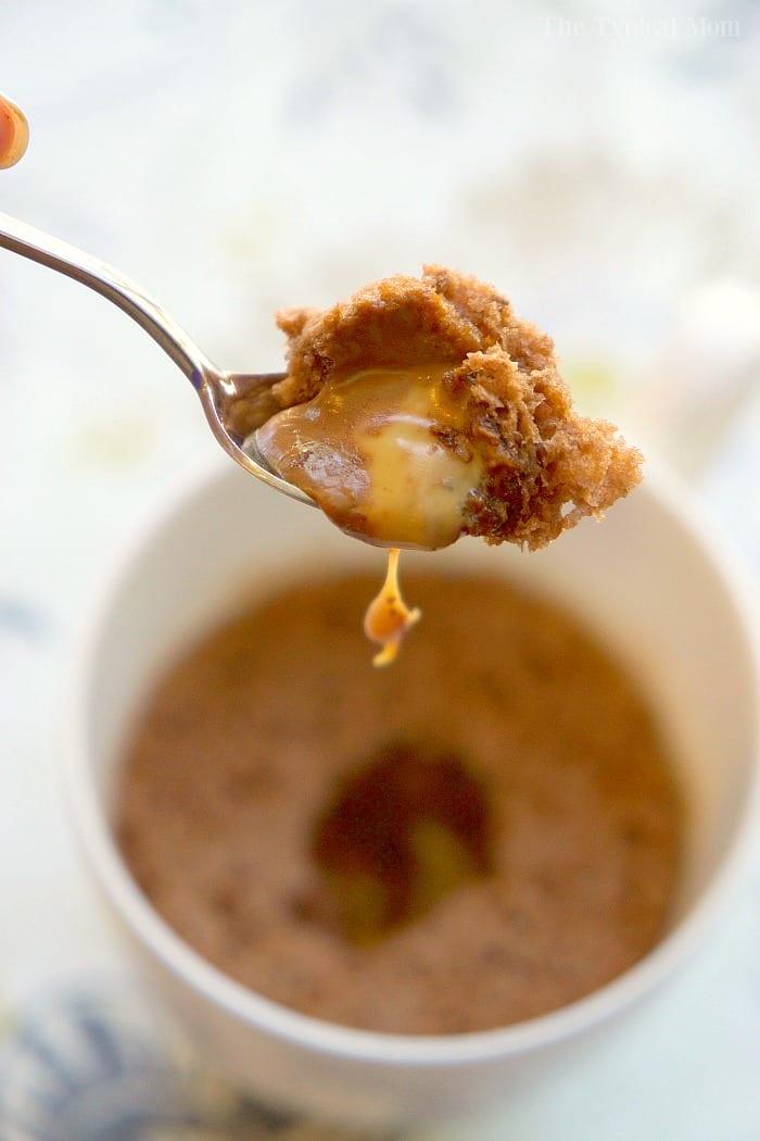 A spoon lifts a bite of molten caramel and chocolate mug cake with a gooey center above a white mug, tempting any dessert lover.