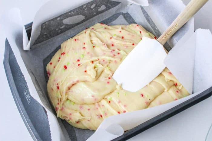 Cookie batter with colorful sprinkles in a parchment-lined pan, being smoothed with a spatula, brings to mind the richness of cookie mix fudge made with condensed milk.