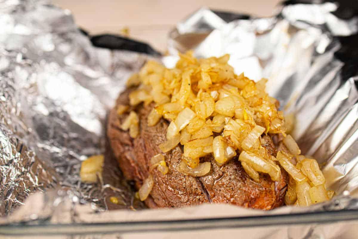 Juicy chuck roll steak cooked to perfection, topped with sautéed onions, and resting on foil in a glass baking dish.