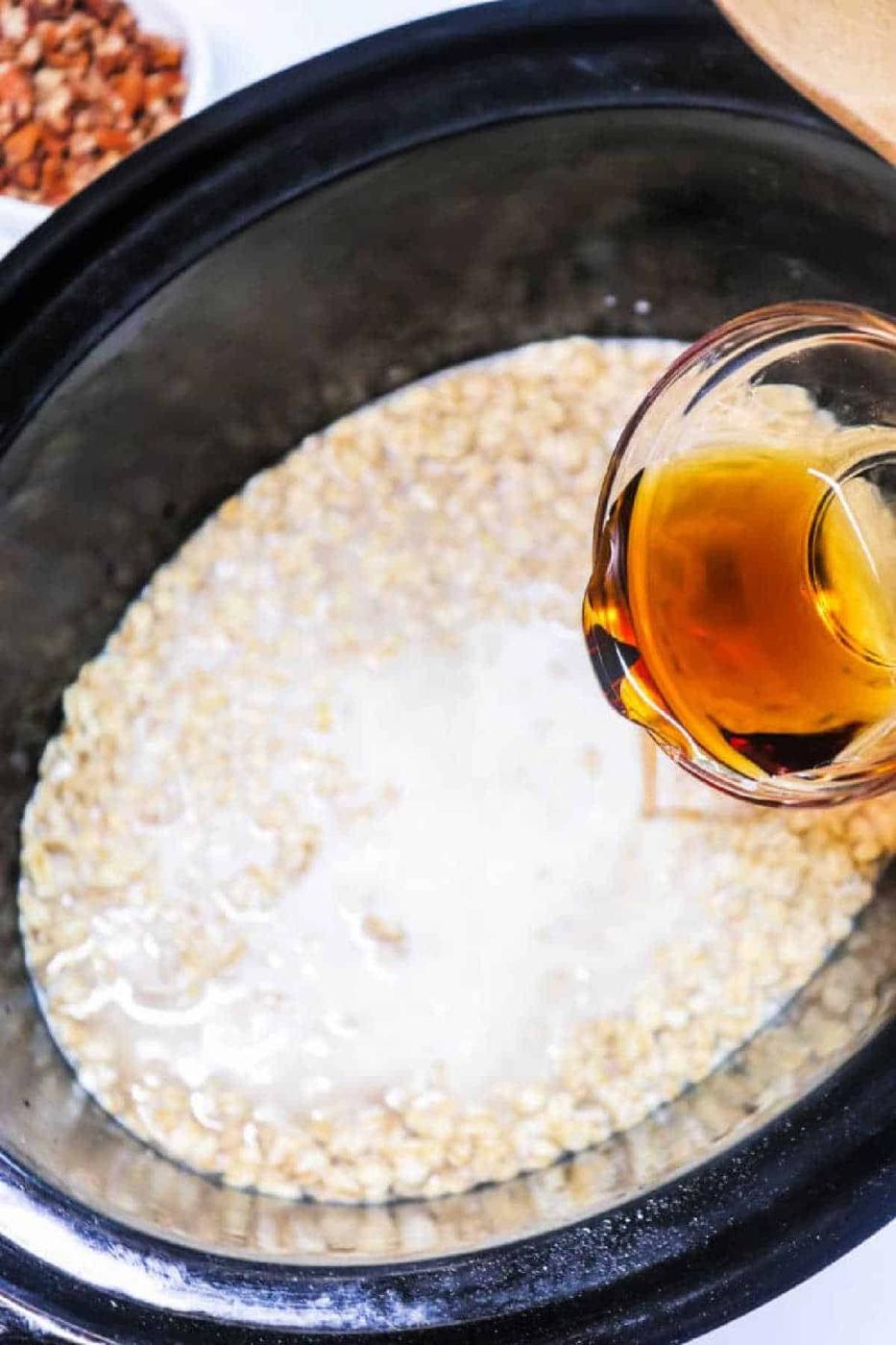 A hand pours a golden liquid into a slow cooker filled with oats and milk, preparing delicious Cinnamon Oatmeal.