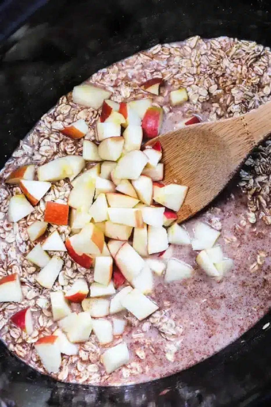Wooden spoon mixing chopped apples, oats, and liquid in a black bowl or slow cooker for delicious cinnamon oatmeal.
