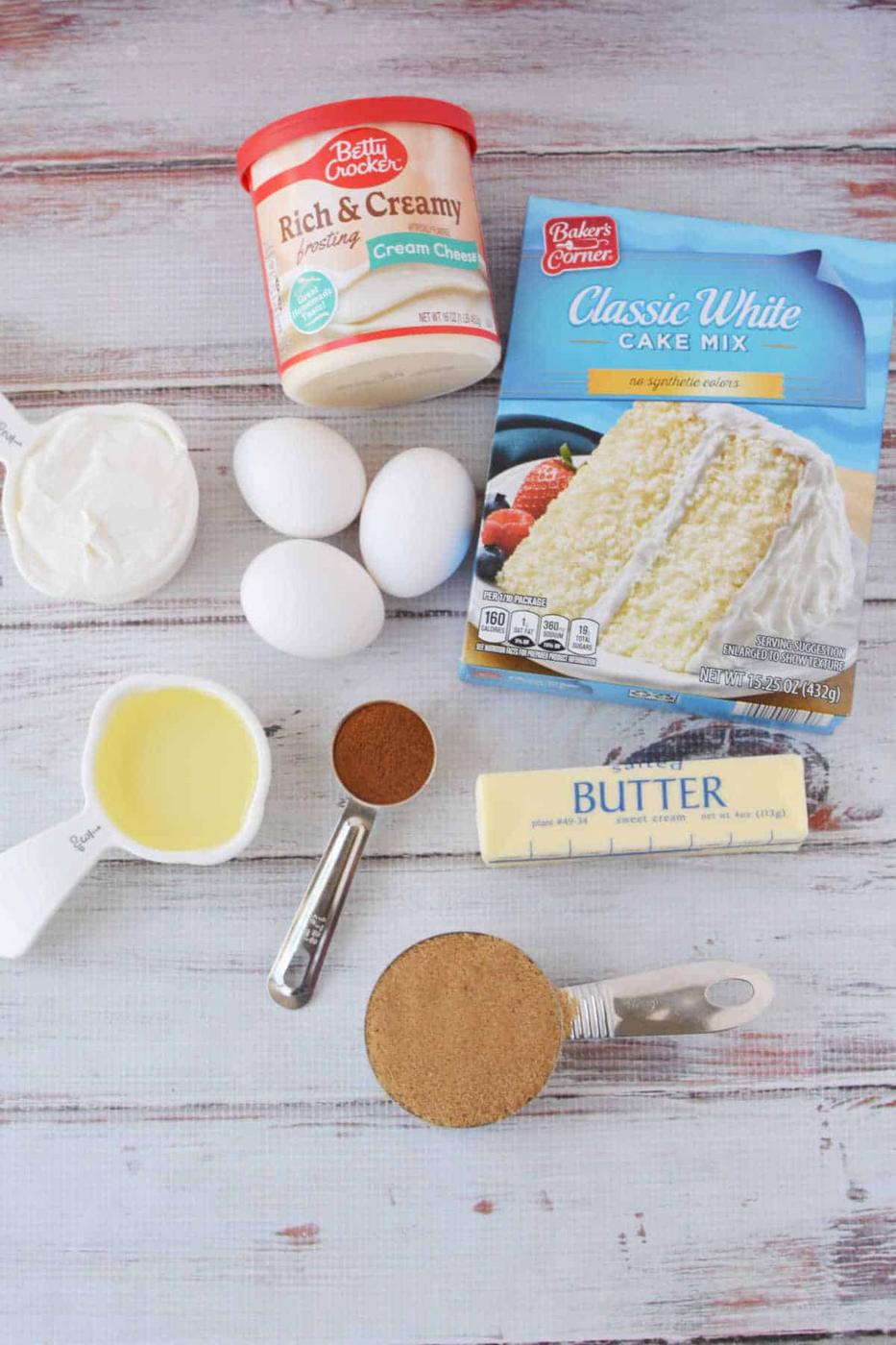 Cake mix, eggs, cream cheese frosting, butter, oil, sour cream, brown sugar, and cinnamon come together to create the perfect cinnamon roll cake on a white table.