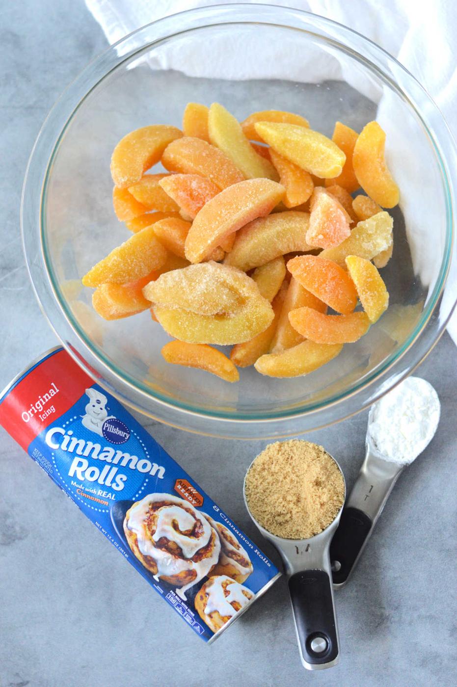 A bowl of frozen sliced peaches, cinnamon rolls can, brown sugar, and flour sit on a gray surface—perfect for making an easy Cinnamon Roll Peach Cobbler.