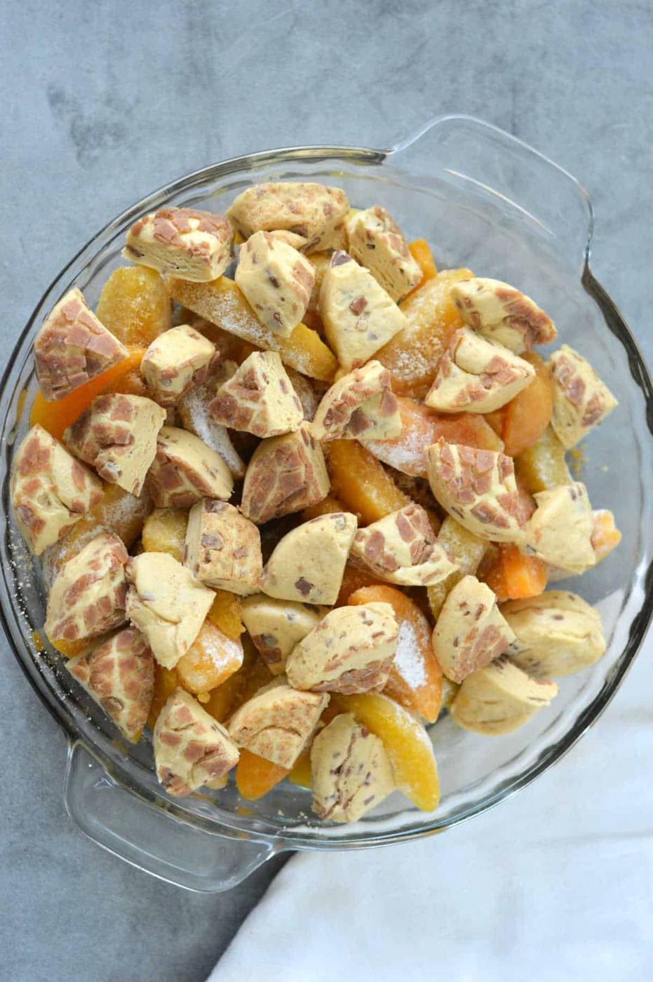 A glass pie dish with sliced peaches and chunks of marbled dough on a gray surface, this Cinnamon Roll Peach Cobbler promises a swirl of sweet, spiced flavor in every bite.