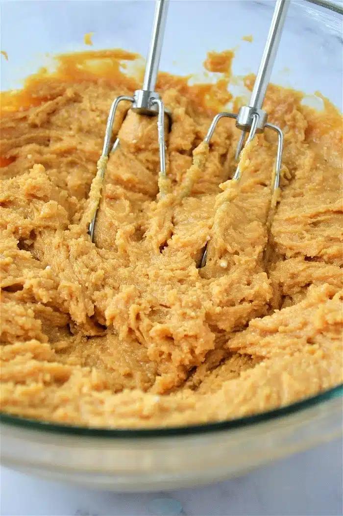 Electric beaters mixing thick cookies made with cake mix dough in a clear glass bowl.