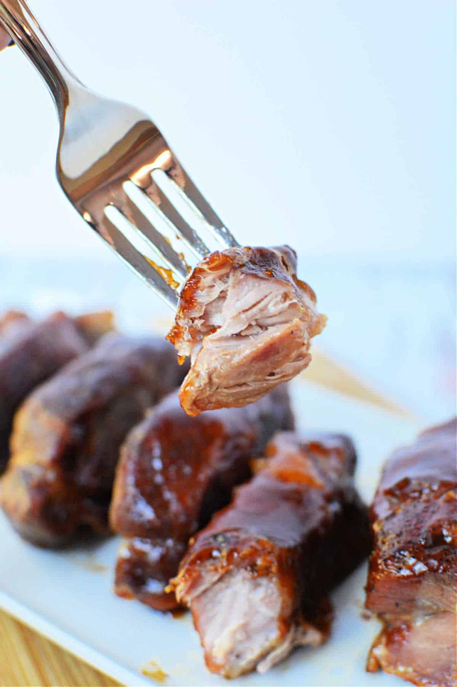 A fork holding a piece of glazed, tender boneless pork ribs above a plate of ribs, showcasing the perfect result of oven cooking.