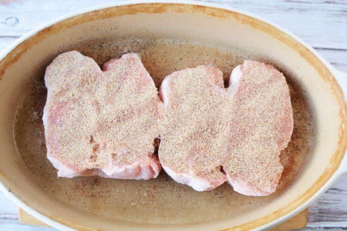 Three uncooked, seasoned frozen pork chops sit in an oval ceramic baking dish on a wooden surface, ready to be transformed into a delicious meal when baked in the oven.